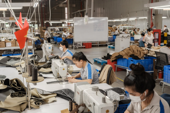 身着蓝条纹灰色短袖工服的工厂工人，正操作缝纫机缝制家具布料。